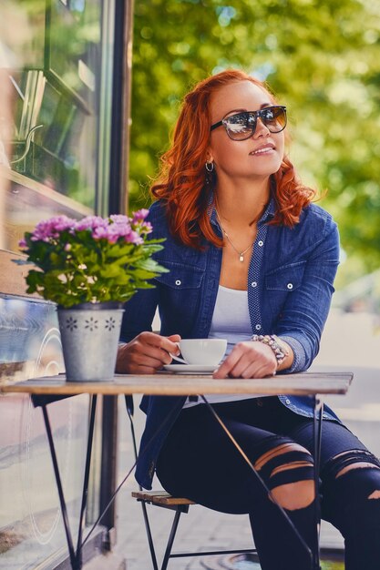 Ritratto di donna rossa in occhiali da sole, beve caffè in un bar in una strada.