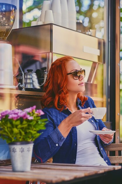 Ritratto di donna rossa in occhiali da sole, beve caffè in un bar in una strada.