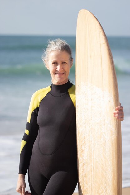Ritratto di donna senior felice che si prepara per il surf in mare. Donna attraente in muta in piedi con la tavola sulla spiaggia sabbiosa vicino al mare e guardando camara. Concetto di sport e salute delle persone anziane