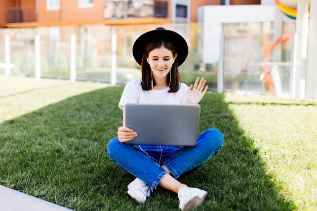 Ritratto di donna socievole che si siede sull'erba verde nel parco con le gambe incrociate durante il giorno d'estate durante l'utilizzo di laptop e auricolari wireless per la videochiamata