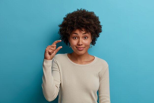 Ritratto di donna soddisfatta con capelli crespi naturali, alza la mano e modella una cosa minuscola, racconta di piccole dimensioni di qualcosa, dice che costa poco sforzo per ottenere il successo, indossa un maglione bianco casual