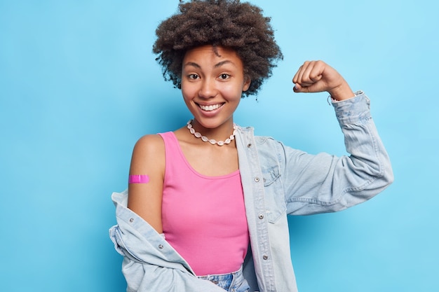 Ritratto di donna soddisfatta dai capelli ricci alza il braccio mostra bicipiti indossa maglietta rosa camicia di jeans collana benda adesiva sulla spalla dopo la vaccinazione si sente bene e protetto incoraggia a vaccinare