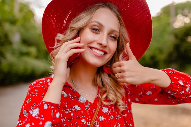 Ritratto di donna sorridente bionda elegante attraente in cappello rosso di paglia e camicetta vestito di moda estiva parlando al telefono emozione gesto positivo
