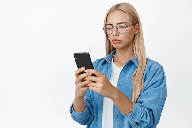 Ritratto di donna triste e cupa con gli occhiali che legge la notifica sul telefono cellulare guardando lo schermo dello smartphone deluso sullo sfondo bianco