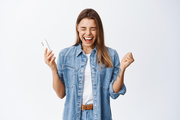 Ritratto di donna urlante felice che vince su smartphone, tiene in mano un telefono cellulare e fa il tifo, celebra la vittoria o il successo in internet, bianco