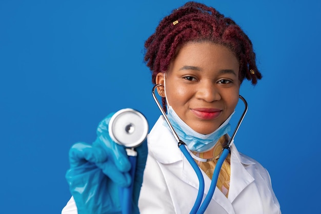 Ritratto di dottoressa africana in camice da laboratorio con maschera facciale e stetoscopio su sfondo blu