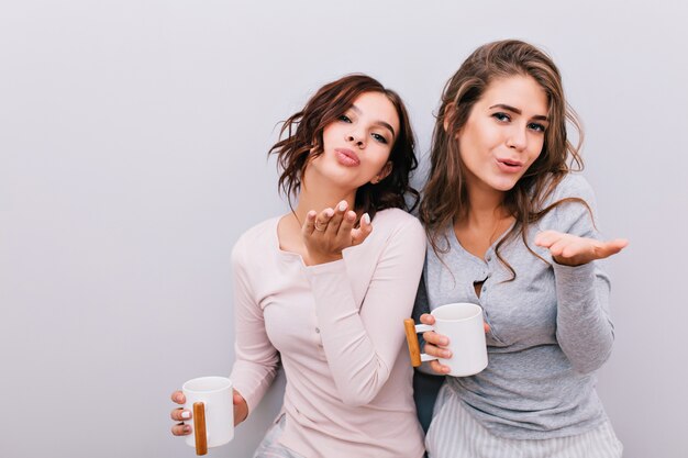 Ritratto di due belle ragazze in pigiama con tazze bianche sul muro grigio. Mandano baci.