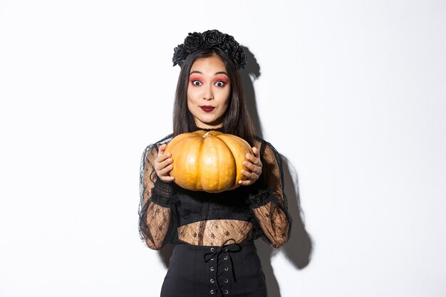 Ritratto di eccitata bella donna asiatica in costume di Halloween, vestita come strega o vedova e tenendo la zucca, in piedi su sfondo bianco.