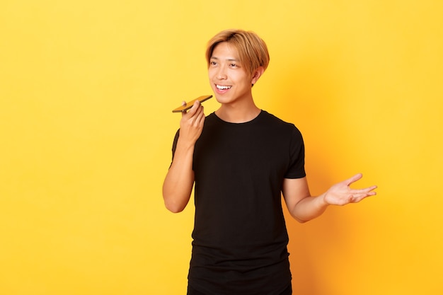Ritratto di elegante bel ragazzo coreano con capelli biondi registrare il messaggio vocale sul cellulare, tenendo lo smartphone vicino alla bocca e parlando, in piedi muro giallo