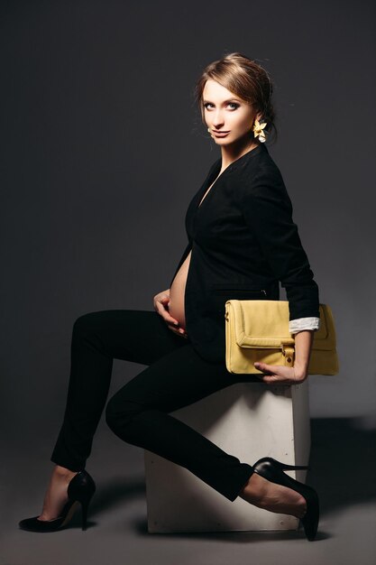 Ritratto di elegante donna incinta che indossa un abito nero con campana incinta guardando sotto la giacca Ragazza bruna con borsa gialla seduta su sfondo nero per studio e guardando di lato