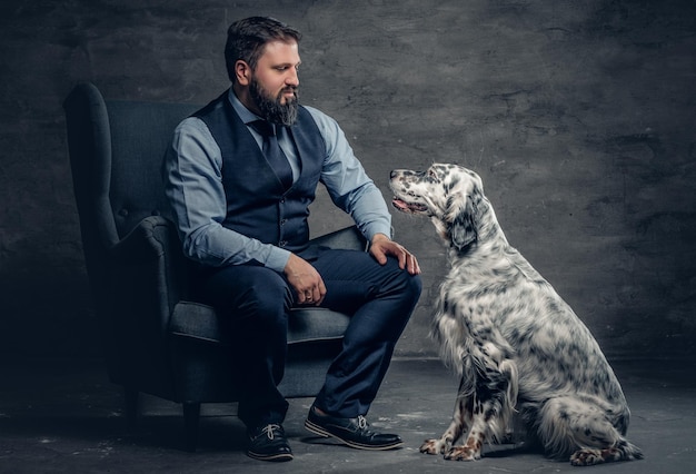 Ritratto di elegante maschio barbuto si siede su una sedia e il cane setter irlandese.