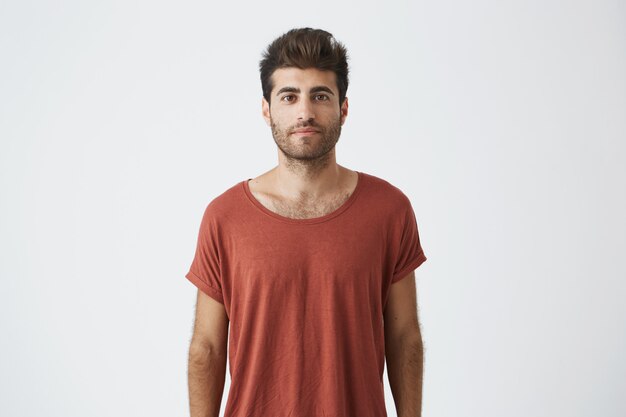 Ritratto di elegante ragazzo barbuto con taglio di capelli alla moda indossando casual t-shirt rossa guardando con i suoi occhi marroni. Giovane uomo bello che ha sguardo soddisfatto. Concetto di persone ed emozioni