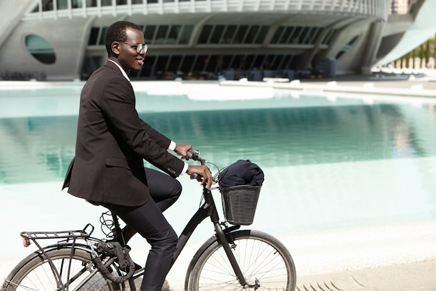 Ritratto di estate all'aperto di giovane impiegato europeo nero bello in occhiali da sole in bicicletta sulla sua bici per lavorare in un ambiente urbano, divertirsi, sentirsi spensierata e rilassata espressione