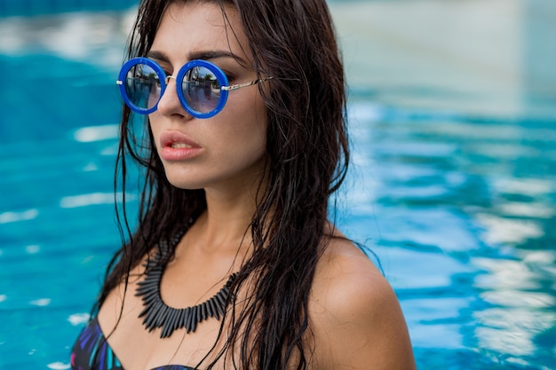 Ritratto di estate della bellissima modella sexy in costume da bagno nero e collana alla moda in posa in piscina. Vacanze e umore tropicale.
