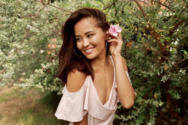 Ritratto di estate della donna asiatica adorabile con il fiore in capelli che posano nel giardino.