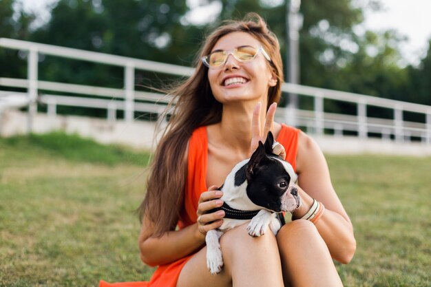 Ritratto di felice bella donna seduta sull'erba nel parco estivo, tenendo il cane boston terrier, sorridente stato d'animo positivo, indossa un abito arancione, stile alla moda, occhiali da sole, giocando con animali domestici