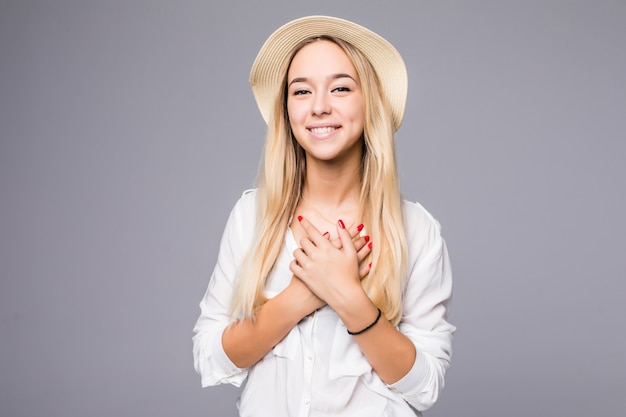 Ritratto di felice bella giovane donna in cappello di paglia si leva in piedi e mise le mani sul suo cuore isolato sopra il muro grigio