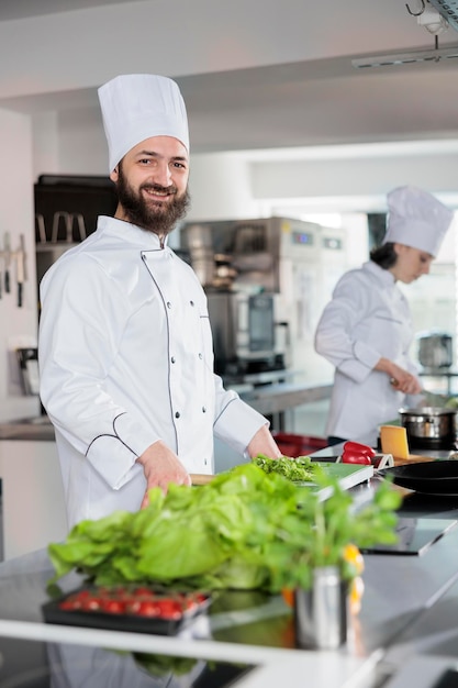 Ritratto di felice capo chef che indossa l'uniforme da cucina, in piedi nella cucina professionale del ristorante mentre sorride alla telecamera. Esperto esperto di cucina gourmet nella preparazione degli ingredienti della ricetta per il piatto da portata.