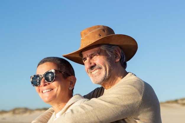 Ritratto di felice coppia senior che trascorre del tempo insieme all'aperto in una giornata di sole. Sorridente uomo dai capelli grigi in cappello da cowboy che abbraccia felice donna dai capelli corti in occhiali da sole. Amore, concetto di svago