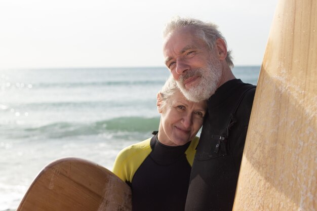 Ritratto di felice coppia senior con tavole da surf vicino al mare. Uomo e donna dai capelli grigi che si abbracciano, guardando la telecamera e la donna che si appoggia al petto dell'uomo. Concetto di relazioni, amore e riposo attivo delle persone anziane