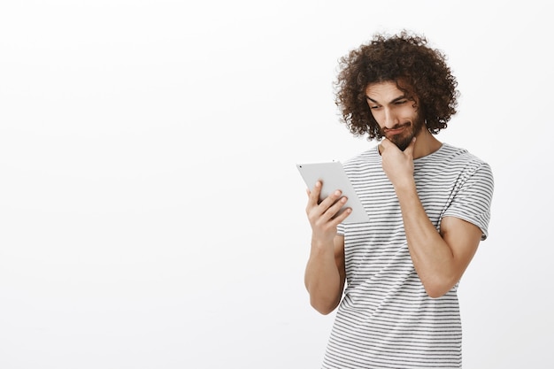 Ritratto di felice curioso attraente ragazzo con barba e taglio di capelli afro, strofinando il mento mentre si guarda la tavoletta digitale, pensando o considerando