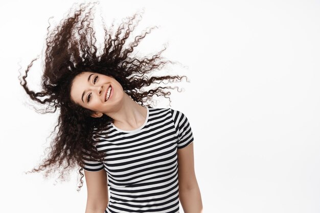 Ritratto di felice donna bruna naturale capovolgere i capelli ricci e sorridente spensierato, guardando allegro, in piedi su sfondo bianco e divertendosi