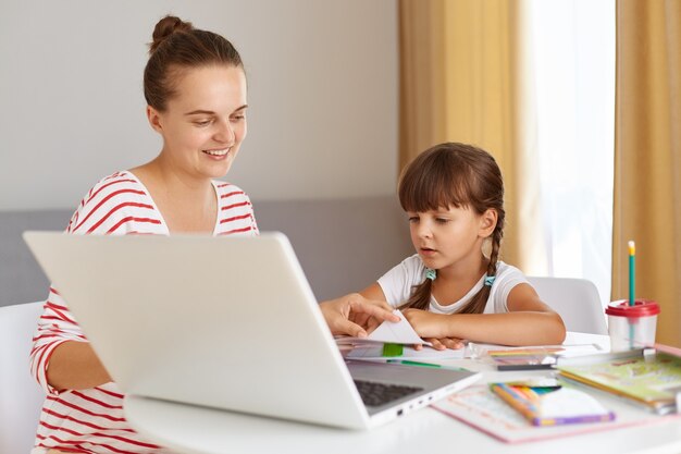 Ritratto di felice donna positiva con figlia che indossa abiti casual, seduta al tavolo contro la finestra in soggiorno, facendo i compiti, madre che aiuta il bambino con la lezione online.