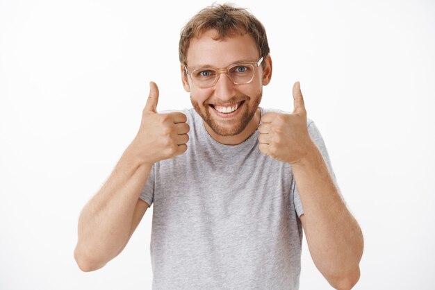 Ritratto di felice felice gioioso manager maschio europeo in maglietta grigia e occhiali sorridente ampiamente mostrando pollice in alto gradimento grande piano o figlia di sostegno sulla concorrenza