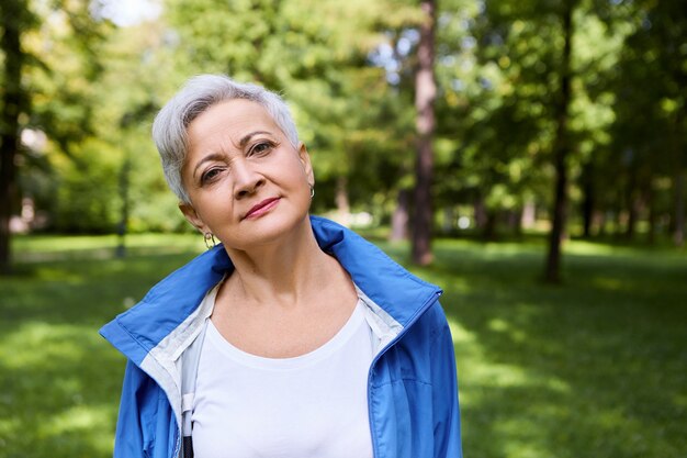 Ritratto di felice femmina caucasica senior con corti capelli grigi rilassante nel parco, con espressione facciale pacifica o premurosa, godersi il tempo da solo nella natura selvaggia, respirando aria fresca e fredda