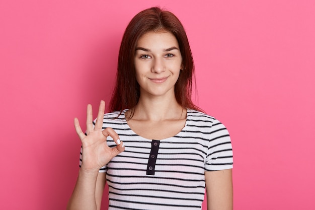 Ritratto di felice giovane ragazza bruna che mostra il gesto giusto con le dita, con un sorriso affascinante, ha ottime notizie, indossa la maglietta a righe.