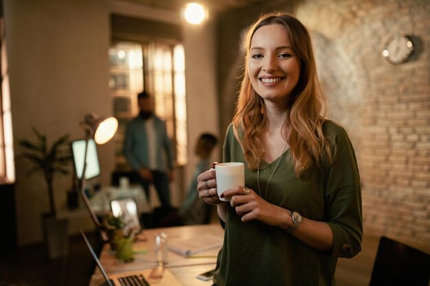 Ritratto di felice imprenditrice che tiene una tazza di caffè mentre lavora fino a tardi in ufficio