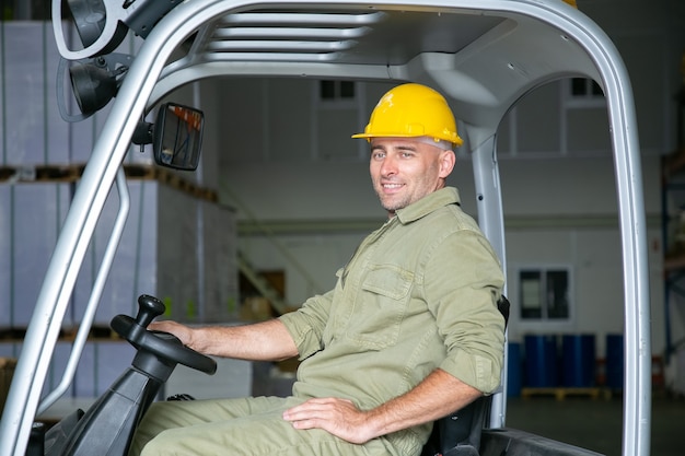 Ritratto di felice lavoratore di magazzino maschio in hardhat guida carrello elevatore in magazzino, tenendo il volante, sorridente, guardando lontano