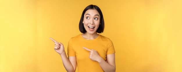 Ritratto di felice ragazza asiatica che punta le dita e guardando a sinistra sorridente stupito controllando il banner promozionale che mostra la pubblicità su sfondo giallo