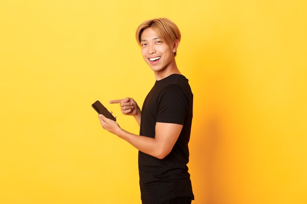 Ritratto di felice sorridente bell'uomo asiatico, in piedi di profilo e puntare il dito sullo smartphone, consiglia l'app, in piedi muro giallo