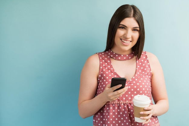 Ritratto di femmina caucasica millenaria che tiene smartphone e tazza di caffè su sfondo turchese