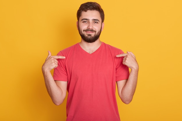 Ritratto di fiducioso bell'uomo caucasico che indossa maglietta casual rossa, in piedi su giallo, che punta sulla sua maglietta