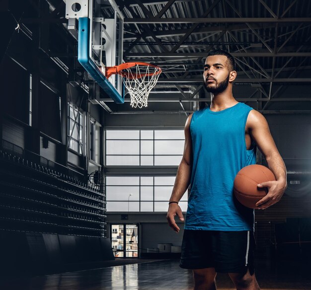 Ritratto di giocatore di basket nero tiene una palla su un cerchio in una sala da basket.