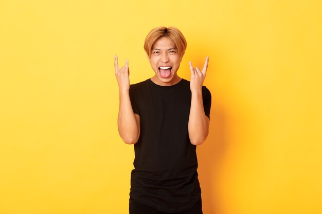 Ritratto di gioioso bel ragazzo biondo asiatico divertendosi, mostrando il gesto rock-n-roll e bastone lingua spensierata, in piedi parete gialla