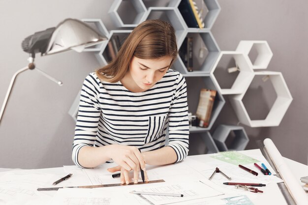 Ritratto di giovane architetto femminile di bell'aspetto serio seduto al suo posto di lavoro, facendo disegni con matita e righello, cercando di non commettere errori nei progetti.