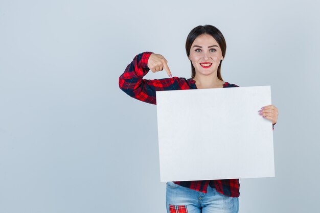 Ritratto di giovane bella donna che punta al bordo bianco in camicia casual, jeans e sembra felice vista frontale