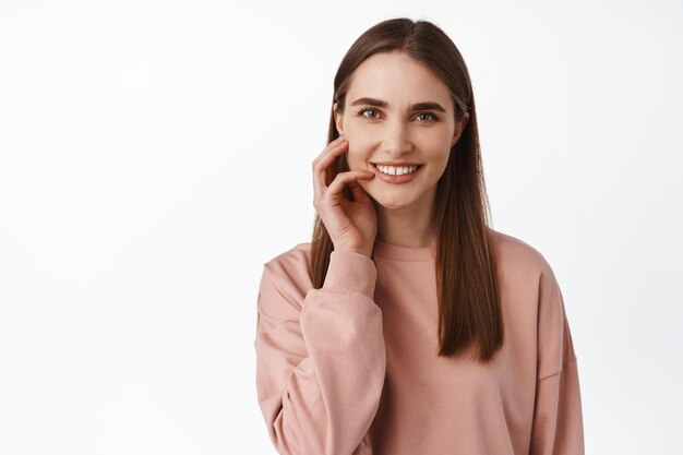 Ritratto di giovane bella donna con pelle pulita perfetta, guancia toccante e sorridente soddisfatto delle condizioni del viso dopo cosmetici per la cura della pelle, sfondo bianco