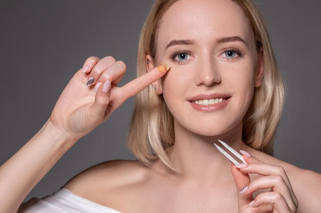 Ritratto di giovane bella donna con trucco naturale e lenti a contatto in mano. Primo piano del modello femminile che tiene una pinzetta bianca per lenti a contatto. Cura degli occhi e stile di vita sano. Salute degli occhi.