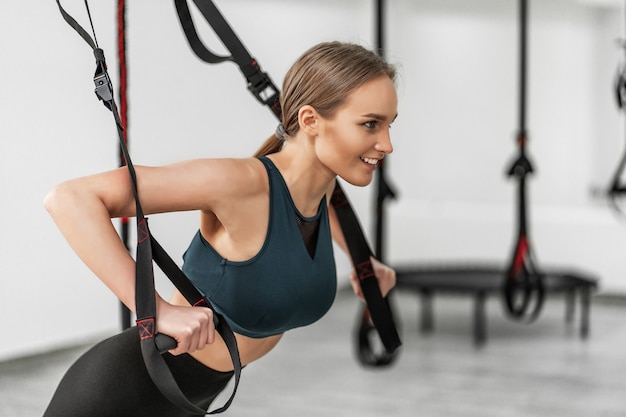 Ritratto di giovane bella donna in braccia di addestramento di abbigliamento sportivo con cinghie di fitness trx in palestra facendo flessioni