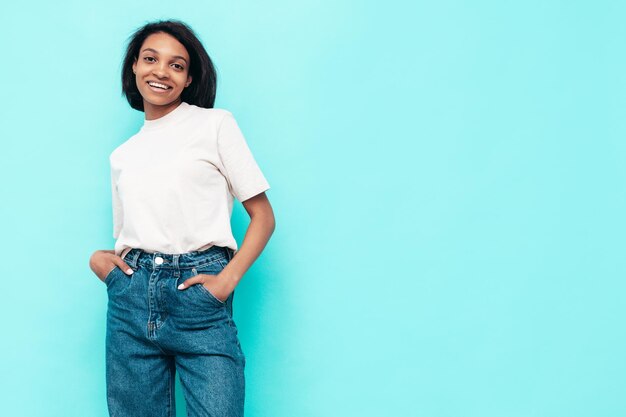 Ritratto di giovane bella donna nera Modello sorridente vestito con jeans estivi Donna sexy spensierata in posa vicino al muro blu in studio Abbronzato e allegro