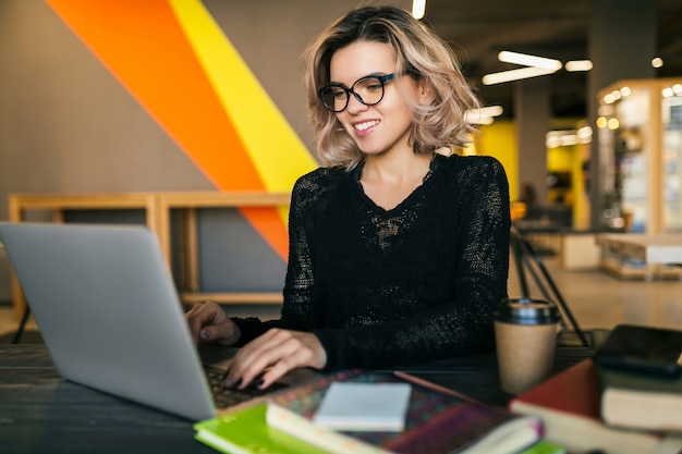 Ritratto di giovane bella donna seduta al tavolo in camicia nera, lavorando sul portatile in ufficio di co-working, con gli occhiali, sorridente, occupato, fiducioso, concentrazione, studente in classe