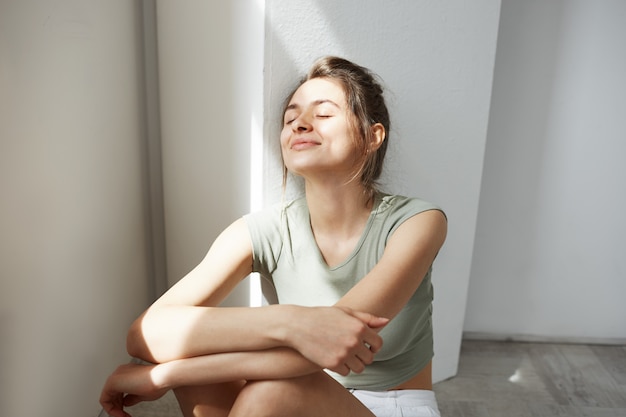 Ritratto di giovane bella donna tenera che sorride con gli occhi chiusi che godono dei sunlights di mattina che si siedono sul pavimento sopra la parete bianca.