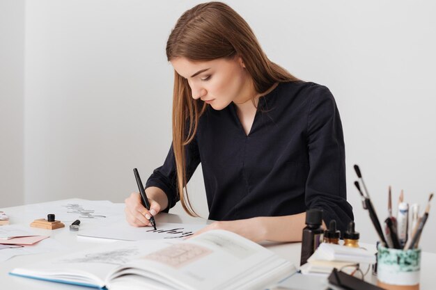 Ritratto di giovane bella signora seduta alla scrivania bianca con un libro aperto mentre scrive note su carta isolata