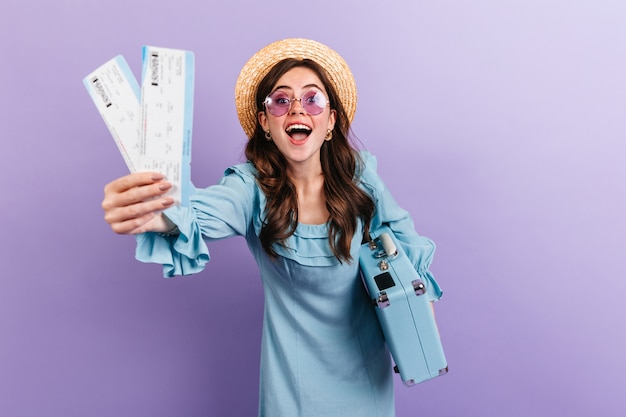 Ritratto di giovane bruna in cappello e occhiali in posa con la valigia sulla parete viola. Donna in vestito blu che si rallegra sinceramente di viaggiare.