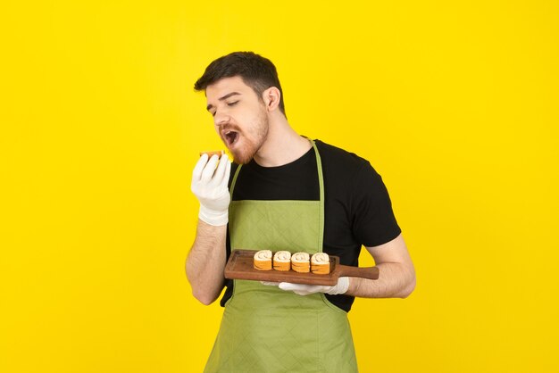 Ritratto di giovane chef che cerca di mordere il rotolo di torta su un giallo.