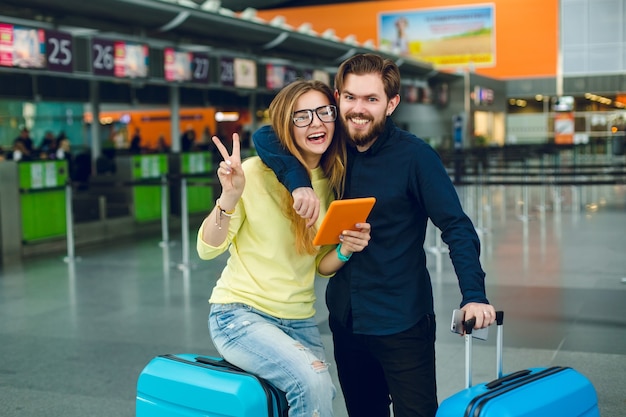 Ritratto di giovane coppia abbracciando in aeroporto. Ha i capelli lunghi, maglione giallo, jeans e tablet. Ha vicino camicia nera, pantaloni e valigia. Stanno sorridendo alla telecamera.